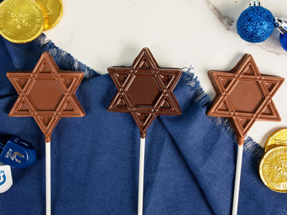 Three milk and dark star pops on a table with other Chanukah decorations.